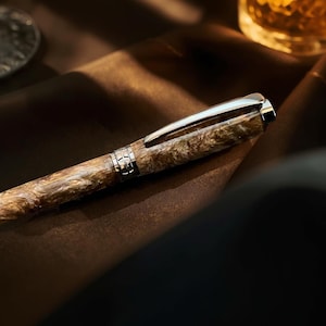 May include: A luxury fountain pen with a marbled brown and beige barrel, silver accents, and a silver clip. The pen is resting on a brown surface, with a blurred glass and liquid in the background.