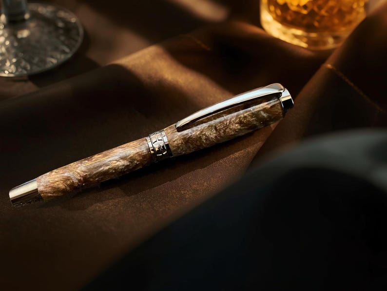 May include: A luxury fountain pen with a marbled brown and beige barrel, silver accents, and a silver clip. The pen is resting on a brown surface, with a blurred glass and liquid in the background.
