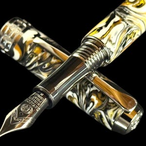 Puede incluir: Una pluma estilogr&aacute;fica de lujo con un dise&ntilde;o veteado en blanco, negro y dorado. La pluma tiene un plum&iacute;n y detalles plateados, con el capuch&oacute;n al lado. La pluma est&aacute; sobre un fondo negro.