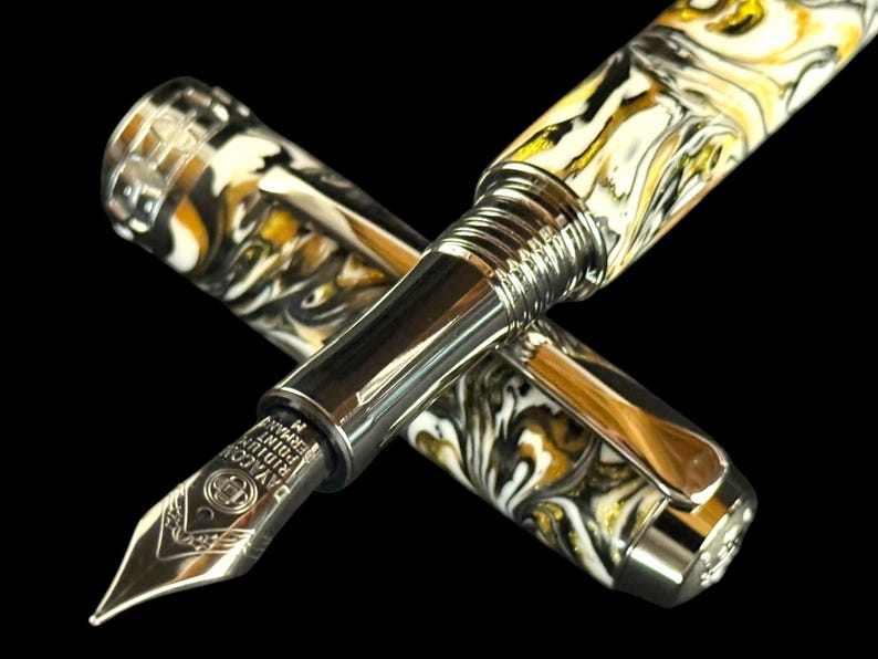 Puede incluir: Una pluma estilogr&aacute;fica de lujo con un dise&ntilde;o veteado en blanco, negro y dorado. La pluma tiene un plum&iacute;n y detalles plateados, con el capuch&oacute;n al lado. La pluma est&aacute; sobre un fondo negro.