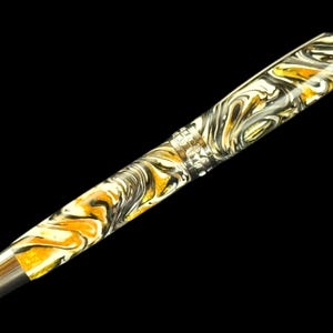 Puede incluir: Una pluma estilogr&aacute;fica de lujo con un patr&oacute;n arremolinado de oro, blanco y negro. La pluma tiene un plum&iacute;n y detalles plateados. El fondo es negro.