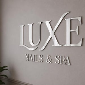 May include: A three-dimensional wall sign featuring the word LUXE in large, reflective silver capital letters, positioned above the smaller text NAILS & SPA in a matching silver finish, mounted on a solid, neutral-toned wall surface.