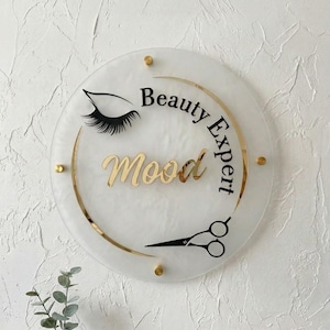 Puede incluir: Un letrero redondo y blanco con detalles dorados y las palabras "Beauty Expert Mood". El letrero presenta una pestaña negra, tijeras y está montado con tornillos dorados. Un artículo decorativo para un salón de belleza.
