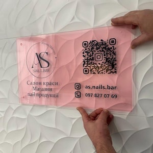 Op de afbeelding: Een helder acrylbord met een roze achtergrond wordt tegen een witte, getextureerde muur gehouden. Het bord bevat de tekst "AS NAILS BAR" in een cirkelvormig logo, samen met een QR-code en contactgegevens. Het bord wordt door twee handen vastgehouden.