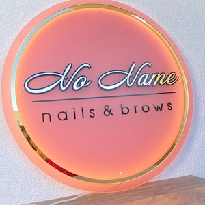 Può includere: Un'insegna rotonda e illuminata con sfondo color corallo e bordatura dorata. L'insegna recita "No Name" in un carattere corsivo, con "nails & brows" sotto in un carattere sans-serif. L'insegna è montata su un muro chiaro.