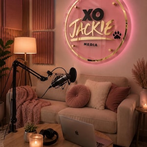 Può includere: Un'insegna da parete circolare con sfondo rosa e bordo dorato, che riporta il testo nero XO JACKIE e MEDIA con un'impronta di zampa nera. L'insegna è illuminata da un anello luminoso a LED rosa ed è montata sopra un divano color beige.