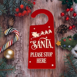 May include: Red wooden door hanger with a white silhouette of Santa Claus in his sleigh with reindeer. The text "Santa, Please Stop Here" is written in white on the door hanger.