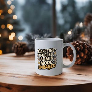 May include: White ceramic mug with the text "Caffeine Fueled Admin Mode: Enraged" in black, yellow, and white. The mug has a curved handle and is sitting on a wooden surface.