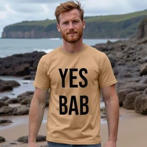 May include: A tan-colored t-shirt with the words "YES BAB" printed in bold black letters. The shirt is short-sleeved and worn by a person standing on a beach with a coastal background.