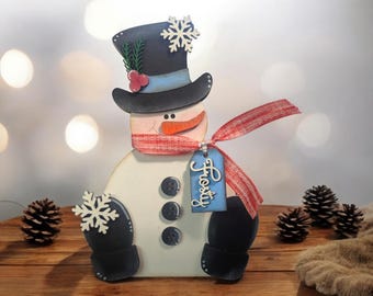 May include: A wooden snowman decoration with a black top hat, a red and white scarf, and black mittens. The snowman has a carrot nose, black button eyes, and a blue tag that says "Frosty". Decorative snowflakes are attached to the hat and mittens.
