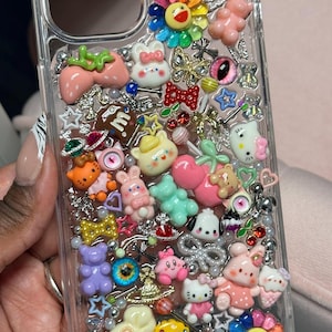 K&ouml;nnte beinhalten: Transparente Handyh&uuml;lle mit einer Auswahl an bunten Charms. Die H&uuml;lle zeigt eine Vielzahl von Designs, darunter Blumen, B&auml;ren, Erdbeeren und Schleifen. Die Charms sind in den Farbt&ouml;nen Rosa, Gelb, Blau und Gr&uuml;n gehalten, mit silbernen Akzenten.