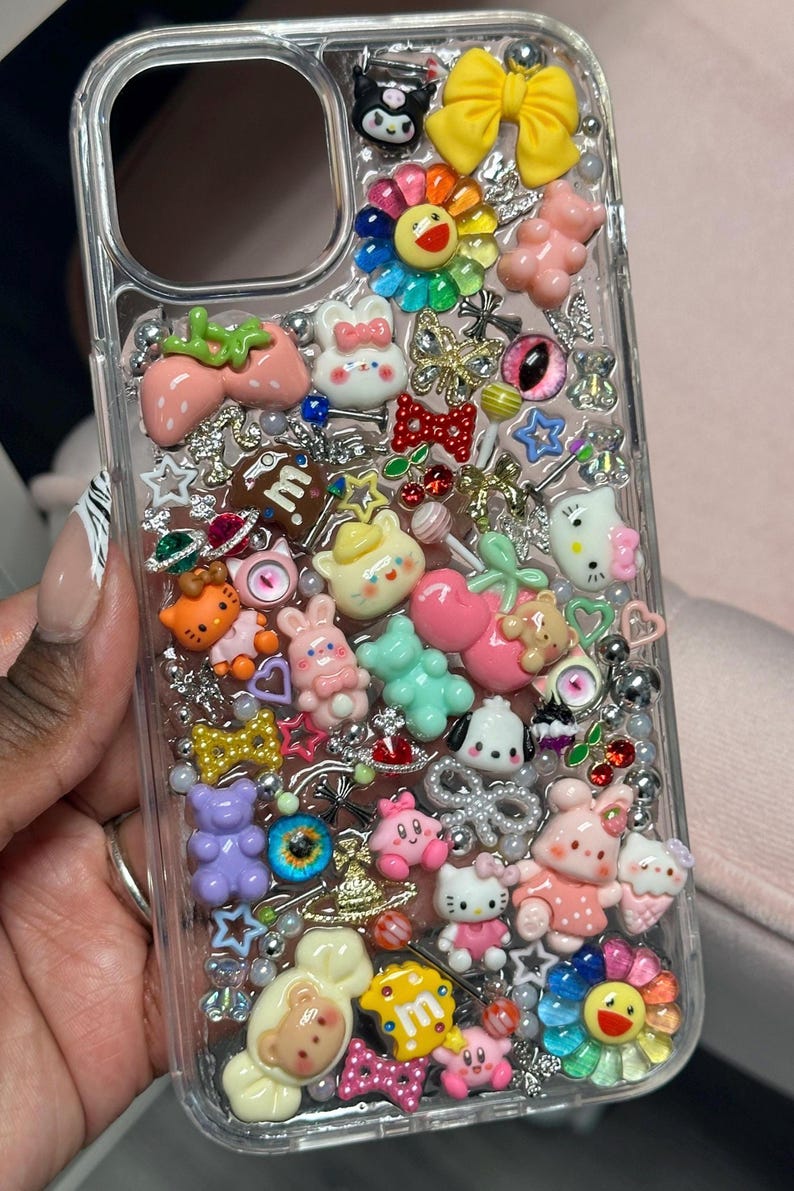 K&ouml;nnte beinhalten: Transparente Handyh&uuml;lle mit einer Auswahl an bunten Charms. Die H&uuml;lle zeigt eine Vielzahl von Designs, darunter Blumen, B&auml;ren, Erdbeeren und Schleifen. Die Charms sind in den Farbt&ouml;nen Rosa, Gelb, Blau und Gr&uuml;n gehalten, mit silbernen Akzenten.