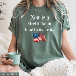 May include: Moss green t-shirt with the text "Now is a Pretti Good time to wake up" in white gothic font. Below the text is a graphic of the American flag. The shirt is a relaxed fit.