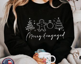May include: Black sweatshirt with a white design of a gingerbread man, a diamond ring, and two Christmas trees. The text "Merry & Engaged" is written below the design.
