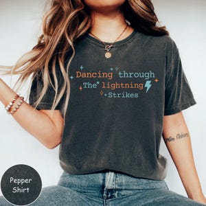 Pode incluir: Camiseta cinza escuro com o texto "Dancing through The lightning Strikes" em laranja, azul e branco. O design inclui um raio e estrelas. A camisa é uma camisa de pimenta.