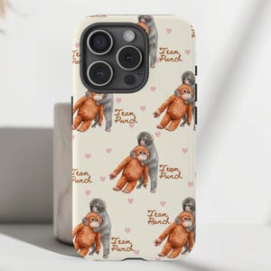 May include: A phone case featuring a repeating pattern of watercolour-style monkeys embracing, with pink hearts and the text "Team Punch". The case is off-white and has a black camera lens.