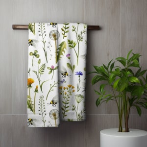 May include: Two white bath towels with a botanical print of wildflowers, bees, and dandelions hanging on a dark brown wooden bar. The towels feature green leaves, yellow, purple, and blue flowers. The towels are hanging in a bathroom.