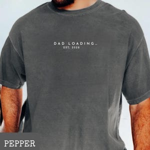 May include: Pepper-colored t-shirt with the text "DAD LOADING... EST. 2026" printed in white. The shirt has a relaxed fit and short sleeves. The shirt is made of a soft, comfortable material.