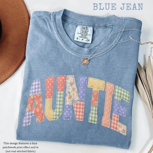 May include: A blue jean-colored t-shirt with the word "AUNTIE" in colorful patchwork letters. The shirt has a crew neck and short sleeves. A silver necklace with a small pendant is draped across the shirt. The shirt is labeled "COMFORT COLORS".
