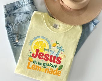 May include: A light yellow t-shirt with the text "Life gave me lemons but my Jesus He be makin' Lemonade" and a lemon graphic. The shirt is folded on top of blue jeans and a beige hat.