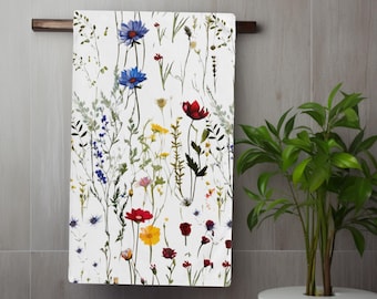 May include: White bath towel with a colorful floral pattern, including blue, red, and yellow flowers, hanging on a wooden bar. The towel is decorated with various wildflowers and greenery, creating a natural, botanical theme.