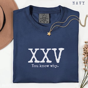 May include: Navy blue t-shirt featuring the text "XXV You know why." A gold-coloured necklace with a star pendant is on the shirt. The shirt has a Comfort Colors tag. A brown hat and flowers are in the background.