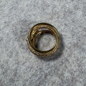 May include: A gold-toned ring featuring a multi-layered, coiled design with a textured, cross-hatched surface pattern. The band consists of overlapping loops that create a continuous, circular shape with a polished finish on the inner edges.
