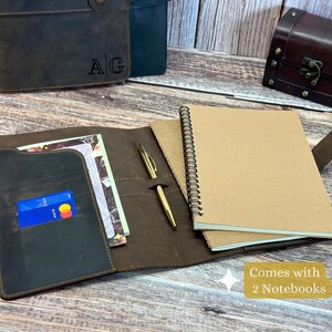 Puede incluir: Una cubierta de cuaderno de cuero marrón con un portaplumas y dos bolsillos. La cubierta tiene las iniciales "A/G" grabadas en relieve en la parte delantera. La cubierta viene con dos cuadernos, uno de los cuales es visible en la imagen.