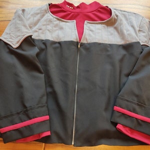 Könnte beinhalten: Eine schwarz-graue Jacke mit rotem Kragen und Manschetten. Die Jacke hat einen silbernen Reißverschluss und gesteppte graue Schulterteile. Die Manschetten und der Kragen haben eine rote Paspel. Die Jacke liegt flach auf einer Holzoberfläche.