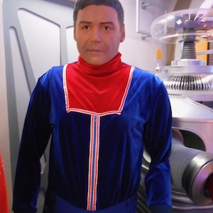 Könnte beinhalten: Eine Schaufensterpuppe in einem blauen Samt-Overall mit rotem Rollkragen und weiß-roten Zierleisten. Die Puppe steht vor einem silbernen Roboter und einer weißen Tür.