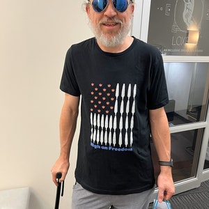 May include: A man wearing blue sunglasses and a black t-shirt with a red, white, and blue American flag design. The shirt has the text "High on Freedom" printed on it. He is holding a black walking stick and a white gift bag.