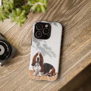 Peut inclure: Coque de téléphone blanche avec une illustration de Basset Hound. Le chien est représenté en marron, noir et blanc, avec de longues oreilles et une expression triste. La coque est posée sur une surface en bois.