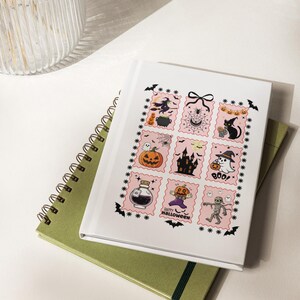 Könnte beinhalten: Ein weißes Notizbuch mit einem Raster aus neun rosa Illustrationen im Briefmarkenstil mit Halloween-Motiven, darunter eine Hexe, eine schwarze Katze, ein Kürbis, ein Spukhaus, ein Zaubertrankfläschchen und eine Mumie, mit dem Text Happy Halloween.