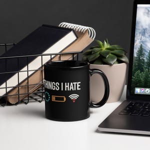 May include: A white ceramic mug with a black handle and the text "But First, Coffee" in a bold, black font. The mug is set against a blurred background, suggesting a cozy setting. The mug appears to be approximately 4 inches tall.