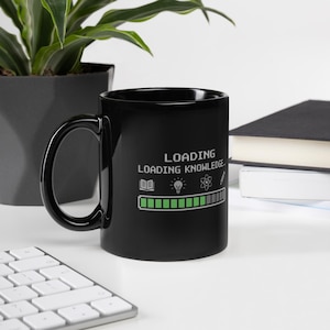 May include: Black ceramic mug with the words "LOADING LOADING KNOWLEDGE" in white. The mug features a progress bar graphic with icons of a book, lightbulb, and atom. The mug has a large, curved handle and is sitting on a white surface.