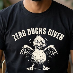 May include: Black t-shirt with the text "ZERO DUCKS GIVEN" above a cartoon duck with an angry expression. The duck has outstretched wings and is standing. The shirt is being worn.