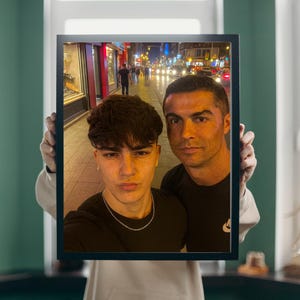 Könnte beinhalten: Gerahmtes Foto mit zwei Männern, die ein Selfie machen. Der Hintergrund zeigt eine Stadtstraße bei Nacht mit Lichtern und Menschen. Beide Männer tragen schwarze Oberteile. Der Rahmen ist schwarz.