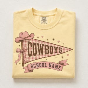 Peut inclure: Un t-shirt jaune pâle avec un motif de fanion à carreaux vichy roses portant le texte COWBOYS. Le design comprend un chapeau de cowboy rose, un nœud, des petites étoiles, des cœurs et une bannière rose en bas avec le texte SCHOOL NAME.