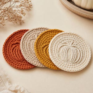 May include: A set of four circular crochet coasters in autumn colors including burnt orange, beige, mustard yellow, and cream. Each coaster features a raised, textured pumpkin design stitched into the center of the round, flat fabric surface.
