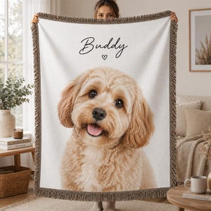 Peut inclure: Un plaid tissé blanc présentant un grand portrait détaillé d'un chien au pelage brun clair et duveteux avec une expression joyeuse. Le nom Buddy est imprimé en écriture noire au-dessus du chien, accompagné d'un petit cœur noir. Le plaid a des franges.