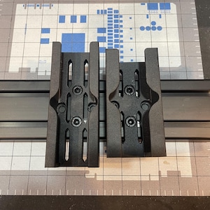 May include: Two black metal brackets with multiple screw holes are mounted on a dark gray rail. The brackets have a symmetrical design with a series of vertical slots. The background is a gray and white grid pattern.