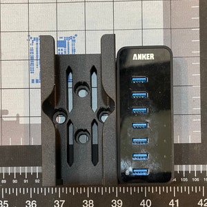 May include: Black Anker USB hub with eight ports and blue inserts, next to a black bracket. The hub has the Anker logo. The bracket has multiple holes and slots. The items are on a grid surface.