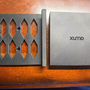 May include: A black metal bracket with diamond-shaped cutouts and a gray square box with the word "xumo" printed on it. The bracket has a series of geometric openings. The box is a solid color.