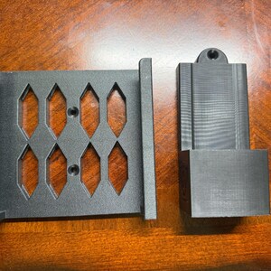 May include: Two dark gray, 3D-printed components. One is a rectangular plate with diamond-shaped cutouts and mounting holes. The other is a rectangular piece with a slot and a circular hole at the top. The items are on a wooden surface.