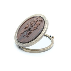 Pode incluir: Espelho compacto ovalado, de estilo antigo, com um design detalhado em madeira entalhada. A madeira escura está inserida numa moldura de metal prateado. O espelho está aberto, revelando a superfície refletora.