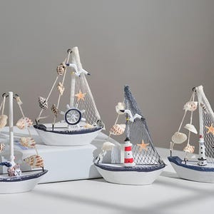 Könnte beinhalten: Vier dekorative Miniatur-Segelboote. Jedes Boot ist weiß mit blauen Verzierungen und verfügt über ein Segel, Muscheln und nautische Elemente wie einen Leuchtturm, ein Steuerrad und Seesterne. Die Boote sind auf einer weißen Oberfläche angeordnet.