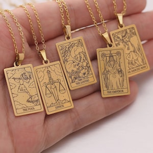 Könnte beinhalten: Goldfarbene Anhängerhalsketten mit Tarotkarten-Designs. Jeder rechteckige Anhänger zeigt eine andere Kartenillustration mit Titeln wie "Der Narr", "Gerechtigkeit", "Die Liebenden", "Der Kaiser" und "Der Gehängte".