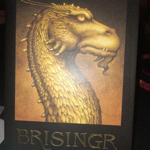 May include: A hardback book with a black cover. It features a golden dragon illustration. The title "BRISINGR" and author Christopher Paolini's name are in gold. The dragon's head is the central image, set against a dark background.