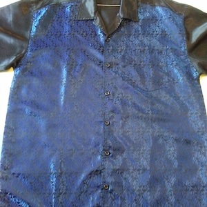 May include: A blue and black patterned button-up shirt with a satin collar and sleeves. The shirt has a black button closure and a pocket on the left side.