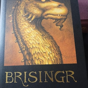 May include: A book titled "BRISINGR" by Christopher Paolini. The cover shows a detailed illustration of a golden dragon's head. The title is in gold lettering against a dark background.